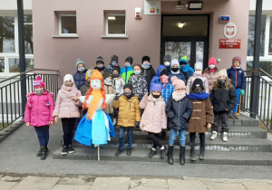 Uczniowie klasy pierwszej w maseczkach, stojący przed szkołą. Wspólnie pozują do zdjęcia klasowego z Marzanną.