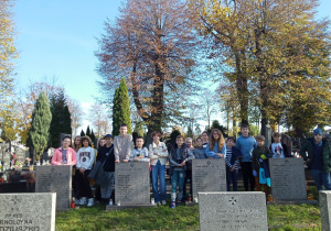 Na zdjęciu uczniowie uczestniczący w porządkowaniu grobów żołnierzy. Przed nimi tablice nagrobkowe z nazwiskami poległych i palące nie przy nich znicze.