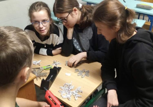 Czworo dzieci, trzy dziewczynki ukłądają wzór śnieżynki, na przeciwko nich siedzi chłopiec z pistoletem na klej i łączy nim wszystkie elementy.