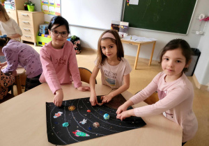 Trzy dziewczynki naklejające i rysujące elementy swojej pracy na czarnym kartonie.