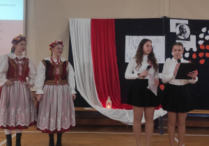 Na tle dekoracji stoją dwie uczennice naszej szkoły, które zapowiadają występ stojących obok nich dziewcząt z zespołu "Anilana"