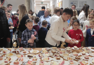 Stół z kawałkami pokrojonych ciast, przed nim zgromadzone dzieci i ich rodzice.