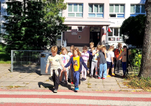 Grupa uczniów stojąca przed jezdnią parami, w tle budynek szkoły .