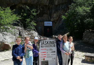 Sześcioro dzieci stojących przy tablicy informacyjnej, przed jaskiniami na kadzielni.