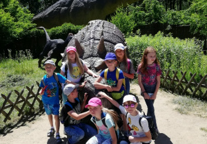 Dzieci pozujące do zdjęcia w Parku Dinozaurów przy jednym z eksponatów. W tle widać inne dinozaury.
