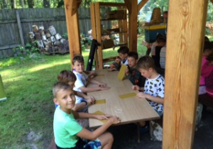Dzieci siedzą przy stole na polanie w oczekiwaniu na zajęcia z lepienia woskowych świec.