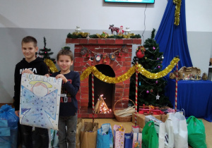 Dwóch uczniów trzyma plakat Aniołkowe Granie w tle paczki z darami oraz dekoracja świąteczna.