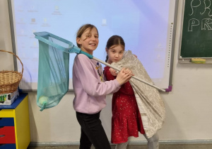 Zdjęcie przedstawia dwie uczennice klasy 3. Jedna z nich trzyma siatkę na motyle, a druga poszewkę, w których to przyniosły swoje zeszyty i przybory szkolne.