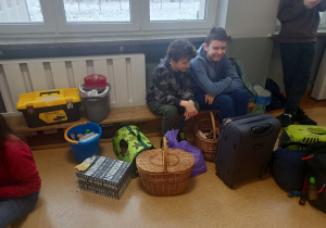 Dwaj chłopcy siedzący na ławce. Przed nimi koszyk piknikowy, skrzynka narzędziowa, transporter dla zwierząt, wiaderko, pudełko kartonowe, walizki, które w dniu dzisiejszym zastąpiły tradycyjne plecaki.