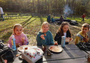 Dzieci siedzące przy stole niedaleko ogniska i jedzące upieczone wcześniej kiełbaski.