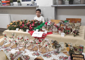 Uczeń klasy 5b, a przed nim na stoisku piękne wianki bożonarodzeniowe, świeczniki ze słoików, pierniki, bombki i inne ozdoby.