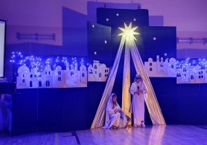 Piękna granatowa dekoracja z zawieszonymi białymi, wyciętymi z papieru budynkami. Na środku zawieszona jest świecąca gwiazda. Przed nią stoją uczniowie odgrywający rolę Maryi i Józefa.