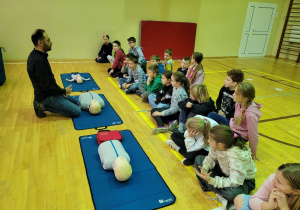 Na zdjęciu ratownik medyczny, tłumaczący dzieciom jakie fantomy leżą przed nim. Na przeciwko mężczyzny, siedzące dzieci.