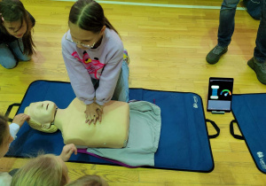 Jedna z uczennic wykonująca masaż serca, obok stojący tablet wskazujący czy dziecko poprawnie wykonuje tę czynność.