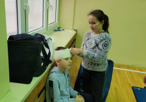 Chłopiec siedzi na ławeczce, koleżanka stoi obok niego i bandażuje jego głowę.