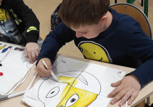 Na zdjęciu chłopiec rysujący na płóciennym worku uśmiechniętą, żółtą buźkę.