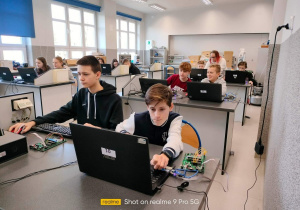 Uczniowie siedzący przed laptopami i podłączonymi do nich podzespołami.