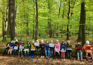 Klasa 1b siedząca w lesie na powalonym pniu drzewa.