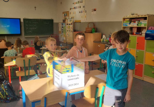 Trzej chłopcy z klasy trzeciej trzymający karty z oddanym głosem nad urną wyborczą.