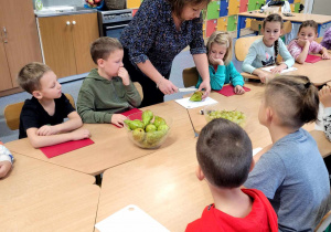 Nauczyciel przekrawający gruszki na pół, dookoła zasłuchana i zapatrzone dzieci. Na stole stoją misy z owocami.