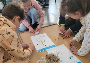 grupka dzieci skupiona przy korkowej tablicy na której jest rysunek konstelacji, sznurek i pinezki. Dzieci tworzą gwiazdozbiory.