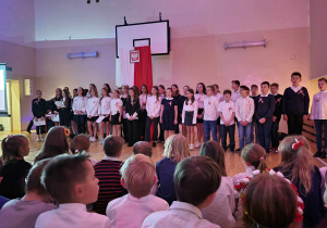 Na zdjęciu chór złożony z uczniów różnych klas oraz kilka osób z widowni wpatrzonych w występujących. W tle biało-czerwony materiał zawieszony na w formie flagi pionowej z godłem na nim.