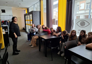 Dzieci siedzące przy stolikach słuchające wyjaśnień jednej z osób prowadzących warsztaty.
