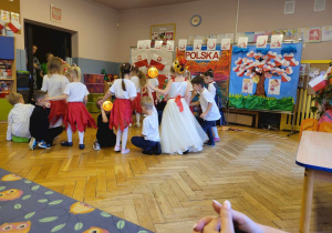 Dzieci z przedszkola tańczące w kole. Chłopcy klęczą na jednym kolanie a dziewczynki trzymając się za palec partnera tańczą wokół niego.
