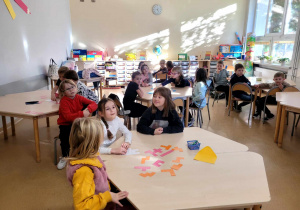 Dzieci podzielone na grupy siedzące przy stolikach, patrzą na nauczycielkę prowadzącą zajęcia i słuchają jej poleceń.