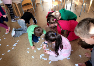 Dzieci szukające karteczek z działaniami, na podłodze leżą karteczki a dookoła kucają dzieci.