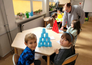 Grupa dzieci siedzącaprzy stoliku na którym ułożone są dwie piramidy z kubeczków plastikowych, obok nich nauczycielka sprawdzająca poprawność wykonanego zadania.