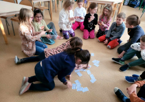 Dzieci siedzące w kręgu, kilkoro losuje karty spośród tych, wyłożonych na środku podłogi.