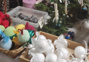 Fragment stoiska na kiermaszu. Na stoliku wykonane przez dzieci kolorowe bombki oklejone włóczką, bombki i szyszki obsypane sztucznym śniegiem.