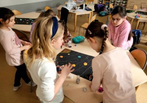 Dziewczynki z klasy pierwszej wokół leżącej na stoliku pracy przedstawiającej kosmos, dopracowujące swoje dzieło.