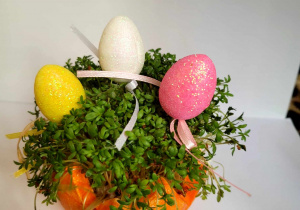 Rzeżucha posiana w połowie styropianowej skorupce jajka pomalowanej na pomarańczowo, ozdobiona kolorowymi brokatowymi jajeczkami.
