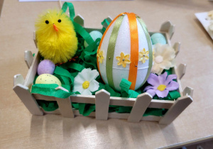 Gotowy drewniany koszyczek wyściełany zieloną bibułą, w nim styropianowe jajko ozdobione tasiemkami i cekinami, kurczaczek, malutkie jajeczka, kwiaty.