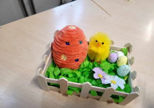 Gotowa dekoracja a w niej: zielona bibuła, żółty kurczak, jajko styropianowe oklejone miękką włóczką, , kwiatki, drobne jajeczka.
