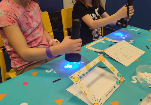 Dziewczynki w okularach ochronnych, trzymające latarki ze światłem ultrafioletowym i święcące na niebieską kartkę na której są ułożone kompozycje z wyciętych białych elementów.