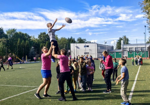 Dwaj zawodnicy klubu rugby podnoszący chłopca , drugi z uczniów rzuca do kolegi piłkę.