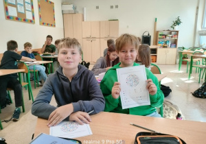 Dwaj uczniowie klasy piątej prezentujący matematyczną kartę pracy. W tle inne dzieci