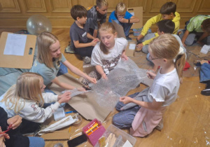 Dziewczynki z klasy 4a tnące folię bąbelkową potrzebną do wykonania pracy podczas warsztatów architektonicznych.