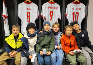 Kilku chłopców z klasy trzeciej w szatni zawodników siedzący na ławkach, za nimi koszulki zawodników reprezentacji Polski