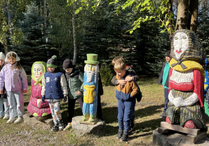 Uczniowie klasy drugiej stojący pomiędzy drewnianymi, ludowymi rzeźbami.