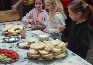 Stół z bułkami, wędliną, warzywami, serem i dżemem, a przy nim dzieci przygotowujące sobie śniadanie.