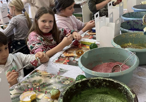 Dzieci siedzące przy stole. przed nimi duże miski z różnymi kolorami brokatów.