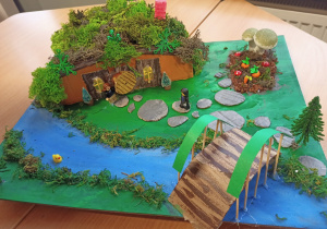 Makieta z ziemianką wykonaną z tektury i mchu, przy niej namalowana rzeka a nad nią mostek.