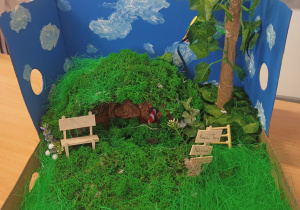 Makieta w pudełku a na niej domek , drzewo, trawa, drogowskazy.