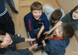 Troje uczniów z nauczycielką, zanurzających ręce w ziemi i tworzących kule kwiatowe.