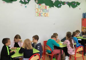 Na zdjęciu dzieci siedzące przy stolikach, pracujące nad jednym z zadań.