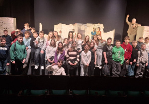 Uczniowie klas piątych pozujący do wspólnego zdjęcia przed sceną, na której znajdują się lalki marionetki i scenografia.
